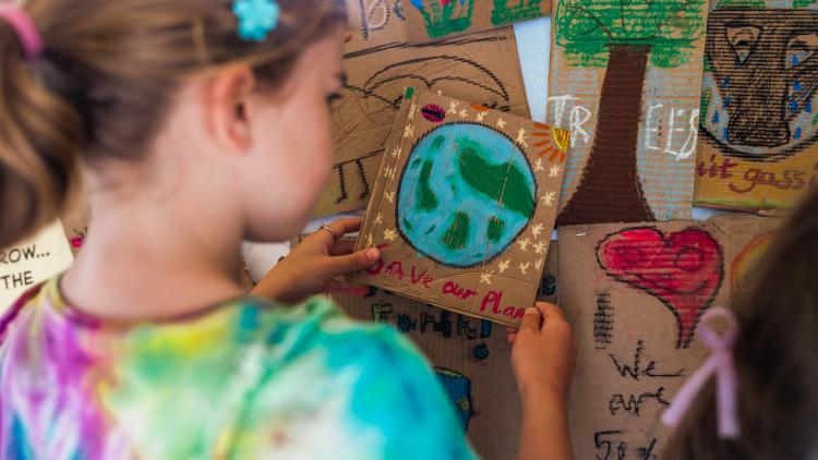 Child placing a handmade cardboard placard with a 'Save our planet' design onto a wall of placards