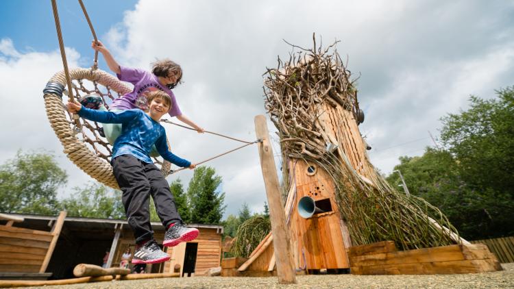 Two children on swing with giant tree climbing frame sculpture in the background
