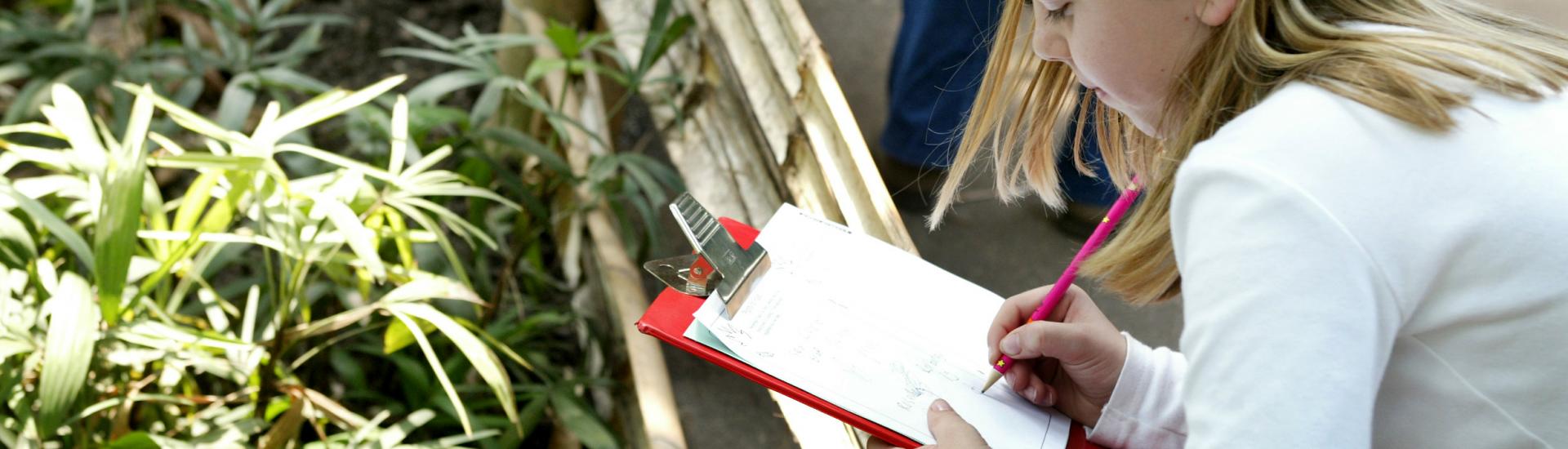 A young girl is looking over a wooden fence at a group of plants and is filling in a sheet of paper on a clipboard