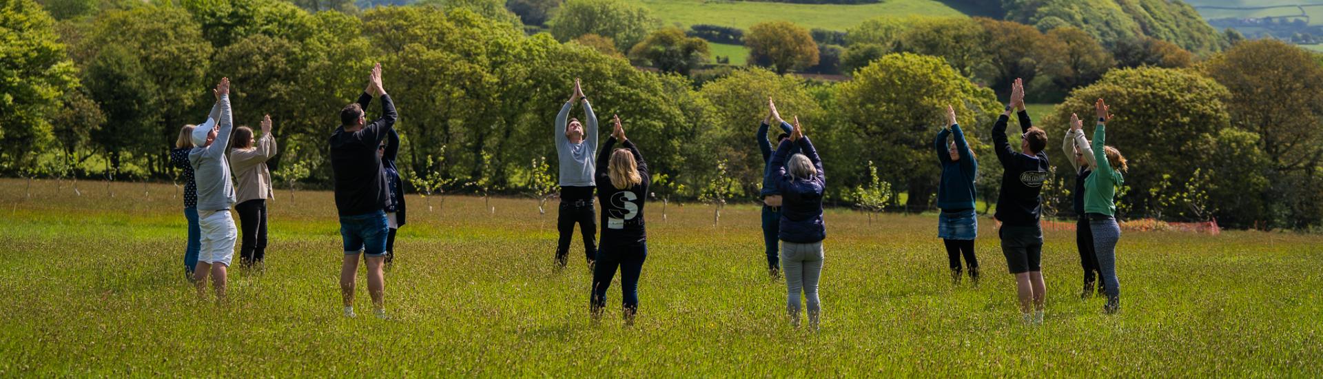 People stood in a circle in a field stretching up with their hands pressed together