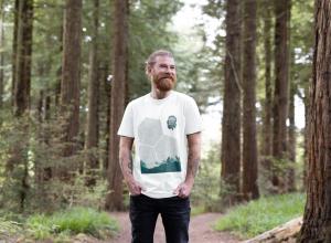 A man modeling the Eden Project marathon tshirt smiles whilst standing in a woodland