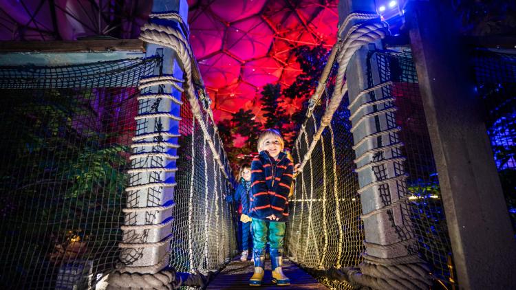 Young boy smiling and crossing a wobbly bridge in the lit up Eden Project Rainforest Biome