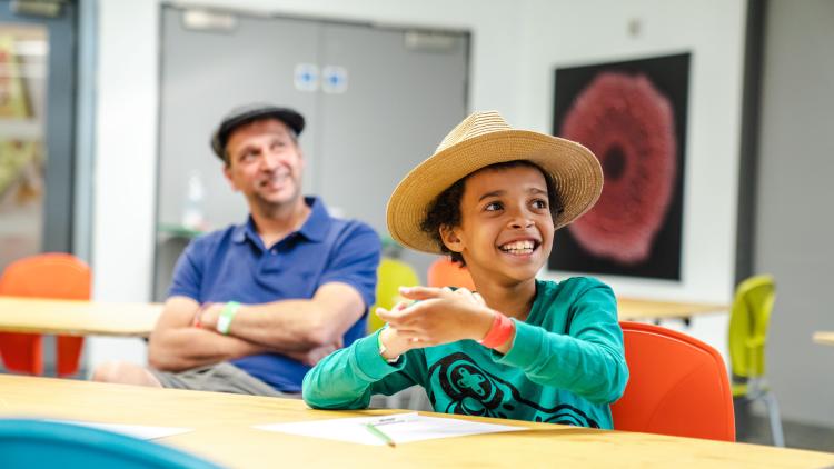 Boy is straw hat smiling doing a writing workshop with his dad smiling in the background