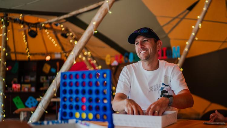 Man in a white t-shirt and black cap smiling and playing Connect 4 in the Tipis 