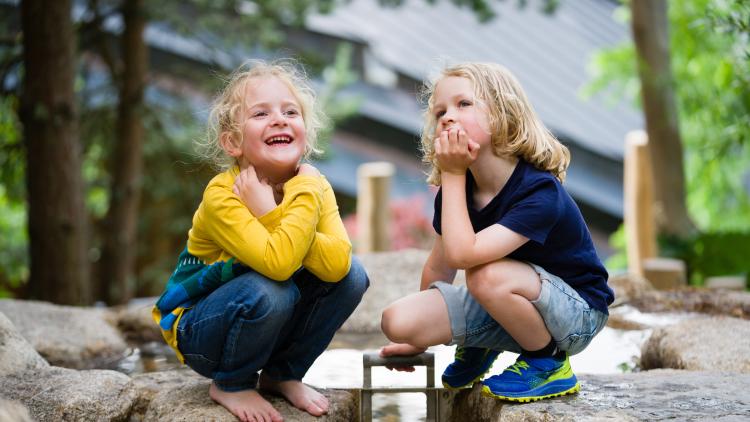 Young girl with blonde hair and in yellow t-shirt and jeans crouching down barefoot playing in the water play course at Nature's Playground, next to a young boy also crouching down
