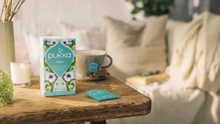 A box of Pukka Herbs 'Relax' blend from their calm range. The packaging is a floral teal and green with flowers on. It's standing on a wooden table with a lit candle