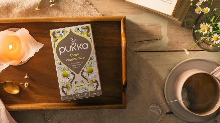 A photograph of Pukka's 'calm' range next to a lit candle and a cup of tea on a wooden table