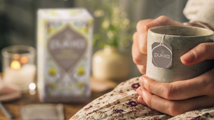 Close up of a hand holding a grey mug with a tag of Pukka tea on the side. In the background, there is a lit candle and a box of Pukka tea