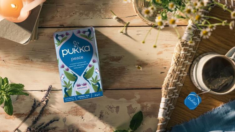A shot of a package of 'peace' tea from Pukka Herbs which is a floral blue and purple print. On the wooden table, there is a lit candle and a cup of tea next to it
