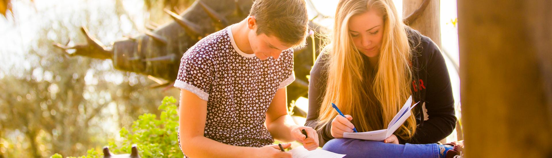 Two school pupils sitting on a wall and writing on clipboards