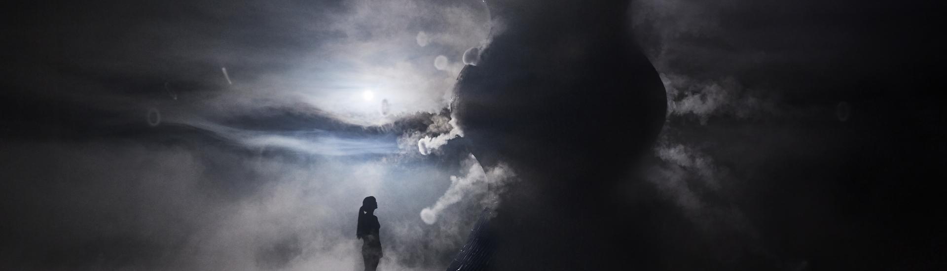 An atmospheric photo which shows a figure standing looking up at the infinity blue sculpture and the vapour surrounding the floor and sculpture 