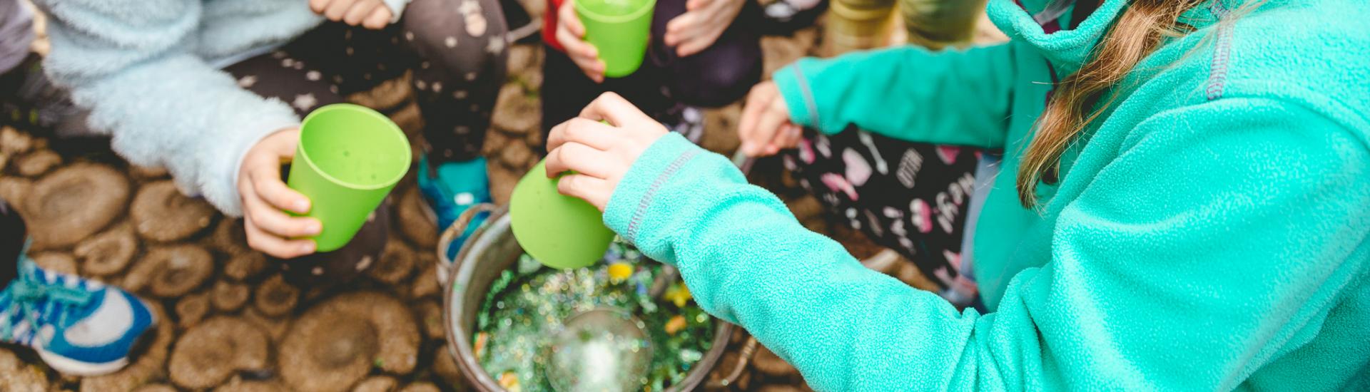 A group of children huddled around a bucket mixing liquids in a bucket and holding cups