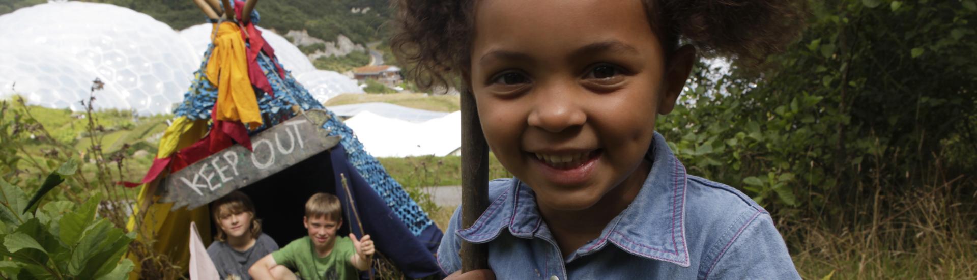 A photograph of two children hiding in a den with the Eden Project in the background and a young girl closer to the camera and holding a stick
