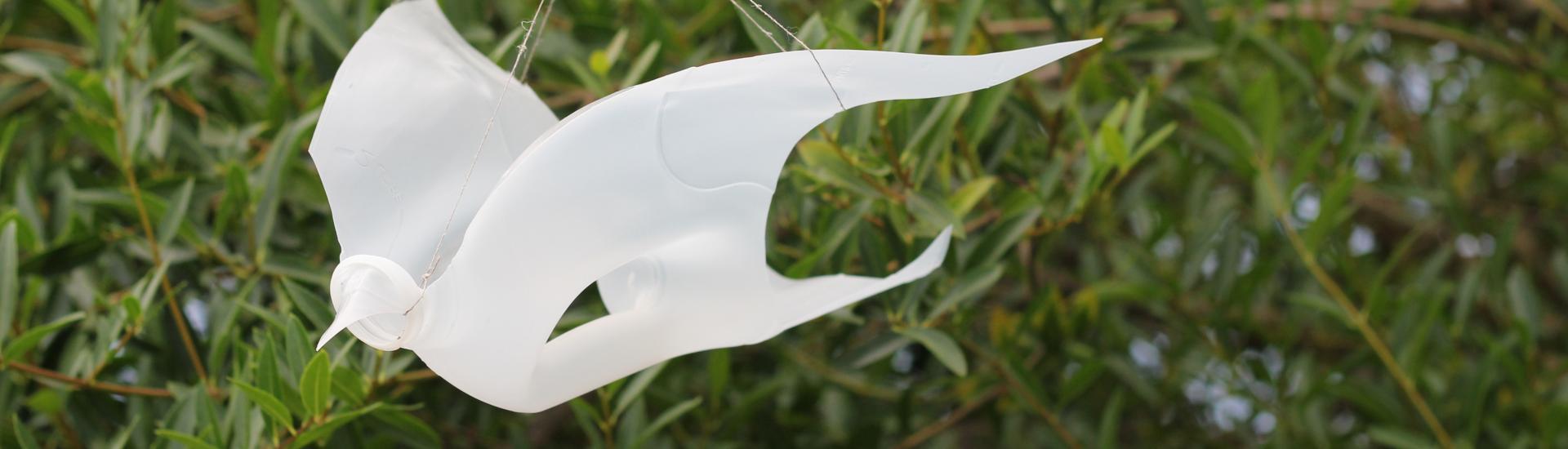 A bird figure made out of a recycled milk bottle hanging mid air with greenery in the background