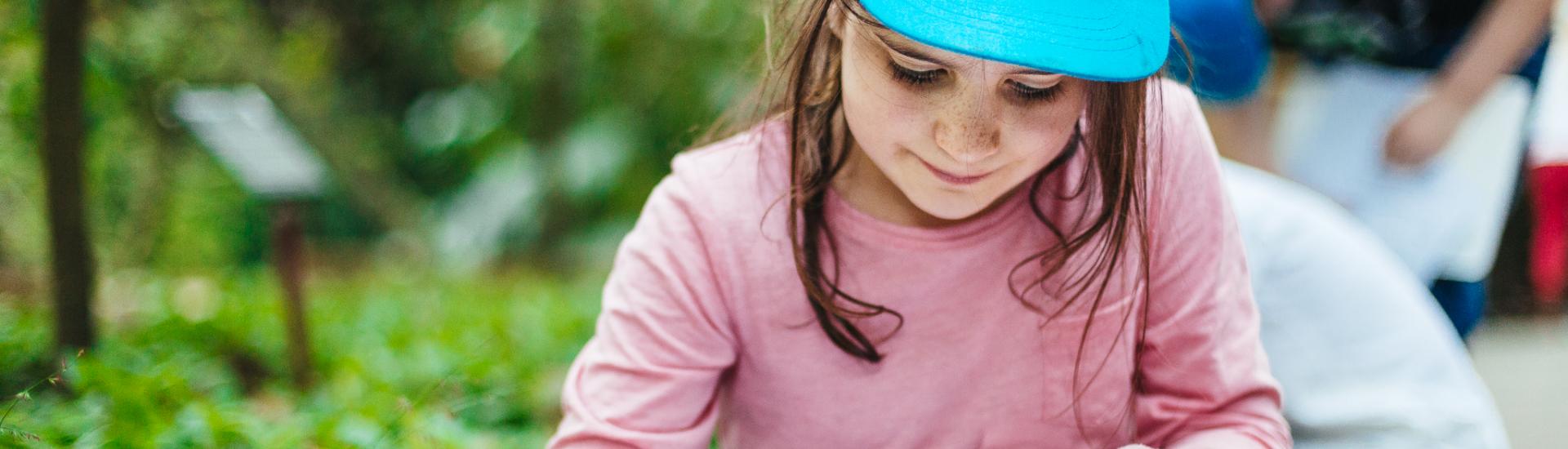 A close up of a young girl with a blue cap on sitting surrounding by greenery and drawing on a piece of paper 