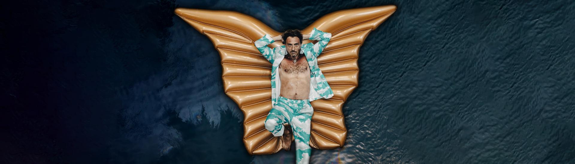 Barechested man wearing a cloud-patterned suit lying on inflatable wings floating on water