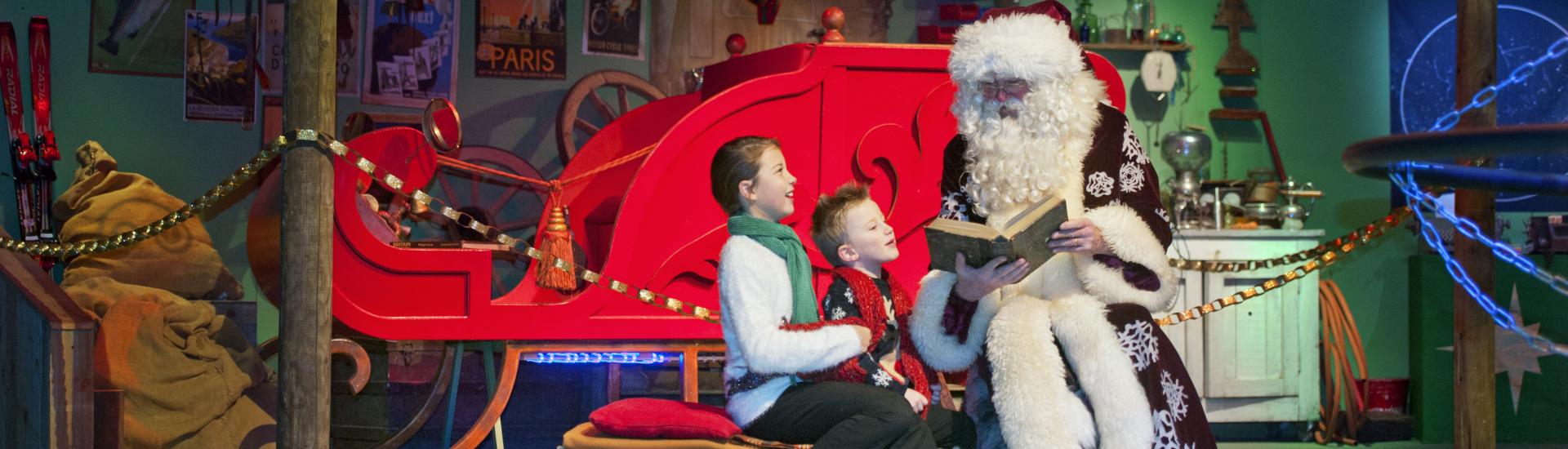 Father Christmas reading to two children in front of a big red sleigh
